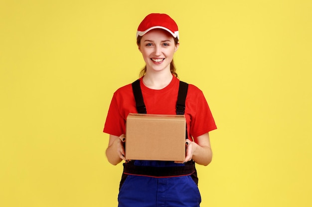 Delighted courier woman standing with cardboard parcel giving it to client looking at camera