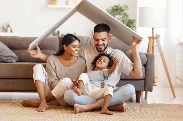 Delighted diverse parents and mixed race son sitting under roof and smiling near sofa in cozy living room at home