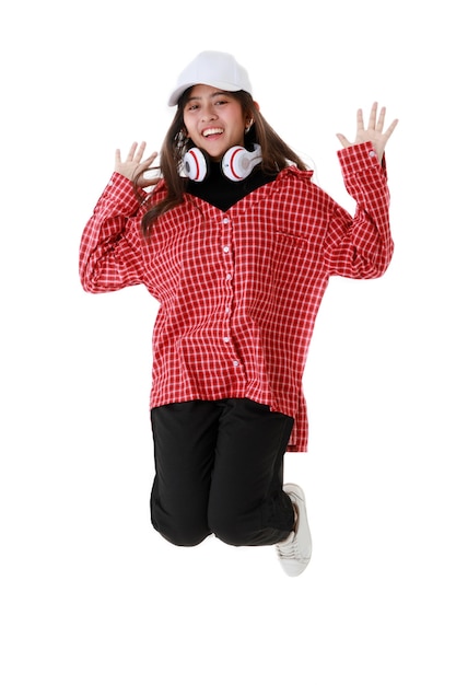 Delighted ethnic female in cool outfit and with headphones on neck in moment of jumping on white background in studio.