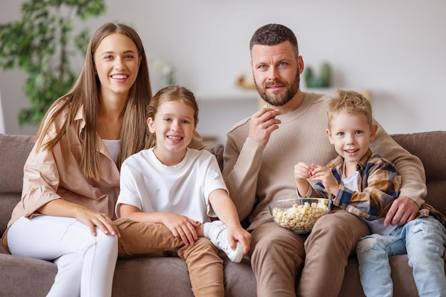 Delighted family parents and kids eating popcorn and laughing while sitting on sofa and watching funny movie at home