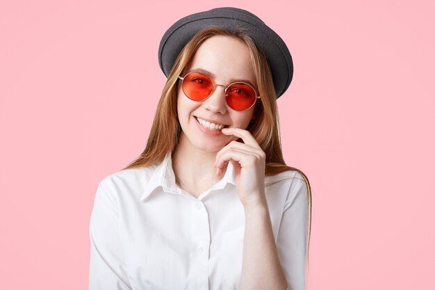 Delighted fashionable hipster girl with broad smile, wears trendy sunglasses and black hat, poses against pink wall, being in good mood after date with boyfriend, isolated over pink wall