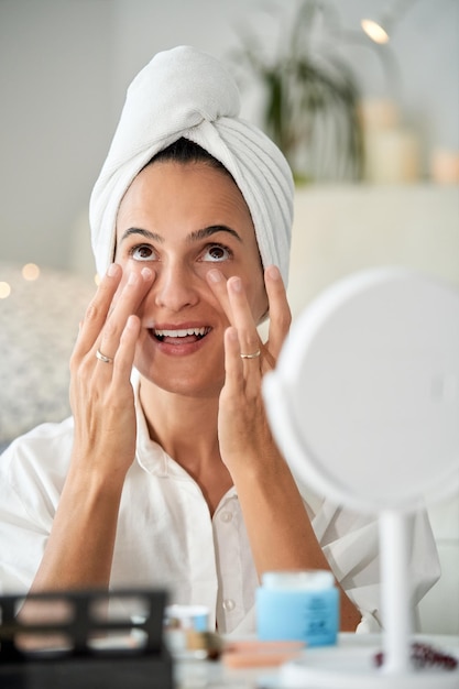 Photo delighted female with soft towel turban on head applying moisturizing facial skin care cream with massage movements on face sitting at table opposite mirror