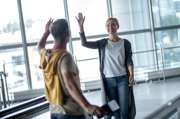 Delighted good-looking blonde woman in casual clothes greeting a male tourist with the boarding ticket