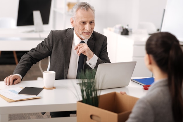 Delighted kind man sitting in the office keeping smile on his face while leaning on the table