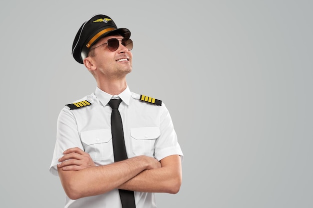 Delighted male pilot in uniform and sunglasses standing with crossed arms on gray background in studio and looking up