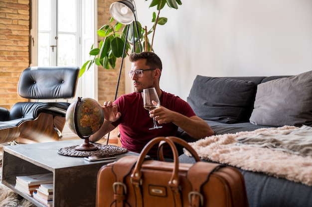 Delighted male traveler choosing country on planet earth globe\
before adventure while sitting in room with suitcase