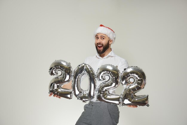 A delighted man with his head up in a red velvet santa hat is\
showing silver balloons in the shape of 2022. a smiling guy with a\
beard at a new year party.