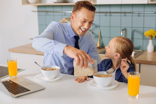 В восторге, милый заботливый отец держит батончик с хлопьями и добавляет его в миску дочери, завтракая с ней.