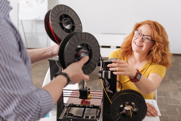 Foto felice positiva donna dai capelli rossi guardando il suo collega e prendendo bobine di filamenti mentre intendeva usarle per la stampa 3d