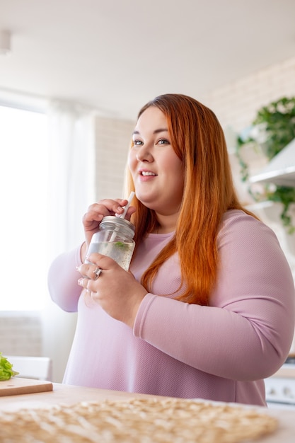 Felice donna positiva in possesso di una bottiglia con frullato in piedi in cucina