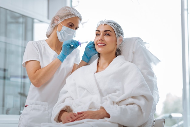 =Delighted positive woman smiling while having a beauty injection