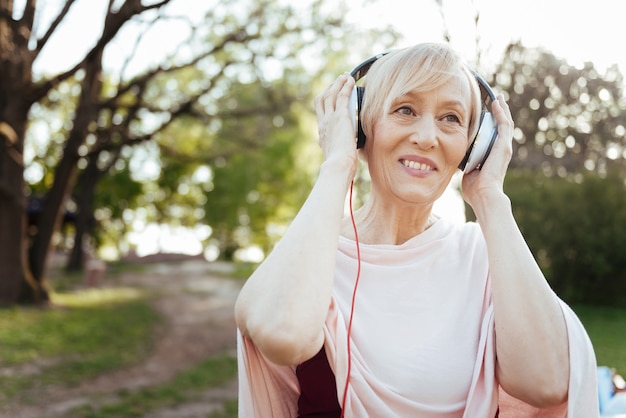 Delighted retired charming woman using headphones while enjoying free time outdoors and listening to the music