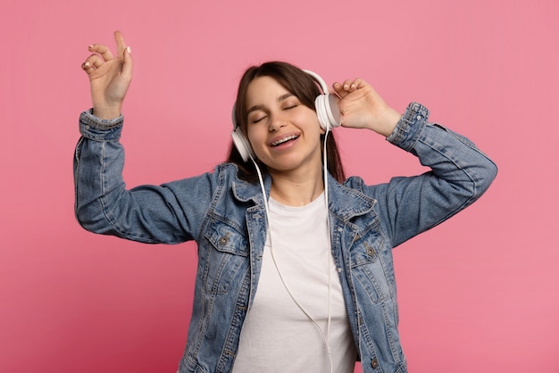 Delighted woman dances in rhythm of melody, closes eyes listens loud song in headphones isolated on pink