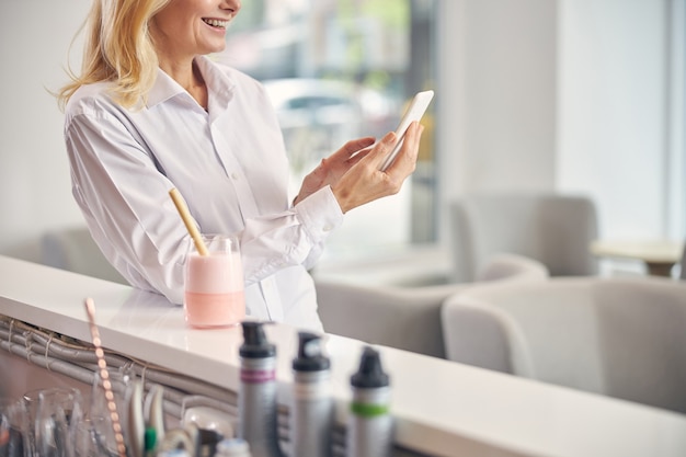 Delighted woman expressing positivity while looking at screen of her gadget, standing near counter