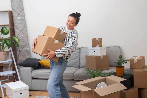 delighted woman moves to her new home living room holding pile of cardboard boxes