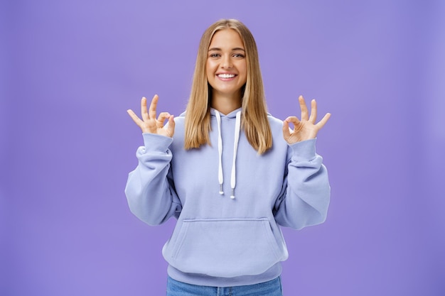 Delighted woman with fair hair showing okay gesture smiling over purple wall.