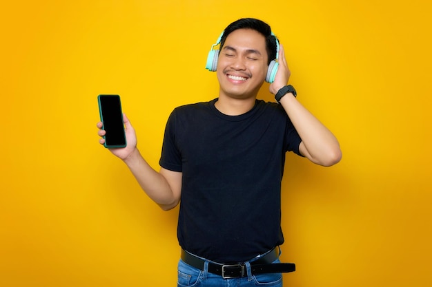 Delighted young asian man in casual tshirt wearing technology wireless headphones for listening to music via mobile phone isolated on yellow background people emotions lifestyle concept