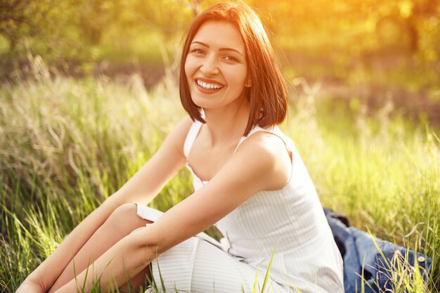 フィールドで晴れた日に緑の芝生に座って笑顔でカメラを見ながら夏のドレスで喜んで若い女性