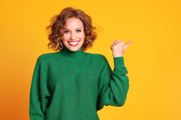 Delighted young female in trendy green sweater smiling for camera and pointing away against yellow background