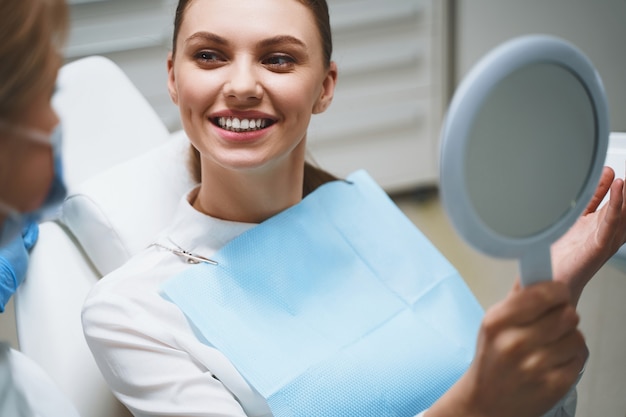 Delighted young girl in dental chair is talking with doctor after treatment and looking in mirror