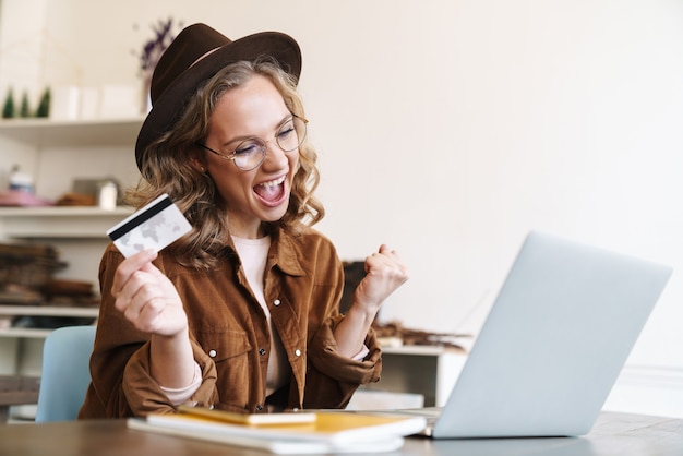 delighted young woman in hat making winner gesture and holding credit card while working with laptop