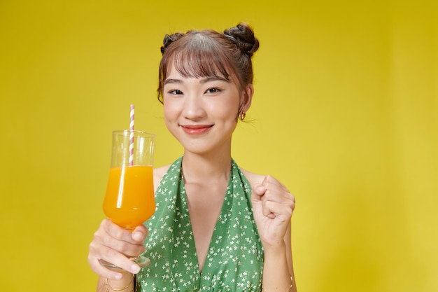 Delightful asian woman drinking orange juice with a straw while standing against yellow