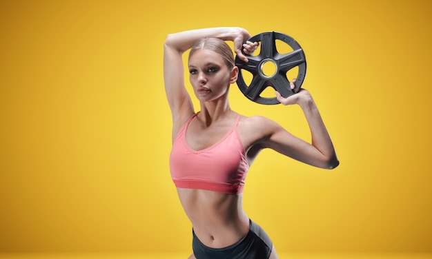 Delightful athlete posing with weights in her hands on a yellow wall.