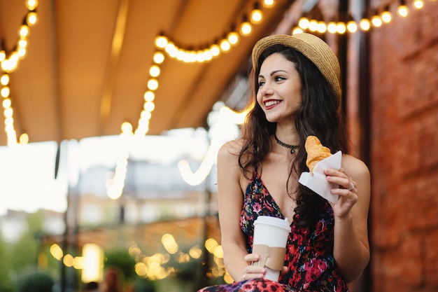 Delightful brunette female with pleasant appearance wearing summer hat and dress holding croissant and takeaway coffee, resting at terrace looking aside with happy expression. People, recreation