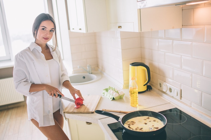 Фотография восхитительная девушка режет помидор на кусочки. она готовит завтрак. есть яйца с грибами, жареные в сковороде на плите. она выглядит и улыбается.