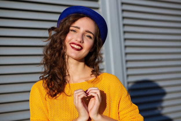 Delightful happy brunette model in yellow knitted sweater and hat making love sign from her hands, space for text