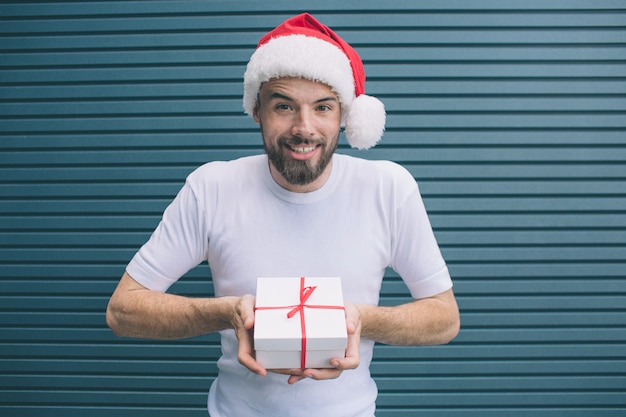 Delightful man in Christmas hat is standing and holding present in white cover. He is looking forward on camera. Guy is smiling. He is very positive. Isolated on striped 