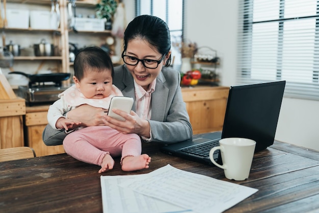 delightful mother in suit cuddling her baby is playing online entertaining nursery rhyme video with phone. happy career woman on table is laughing and showing fun pictures to her child on phone.