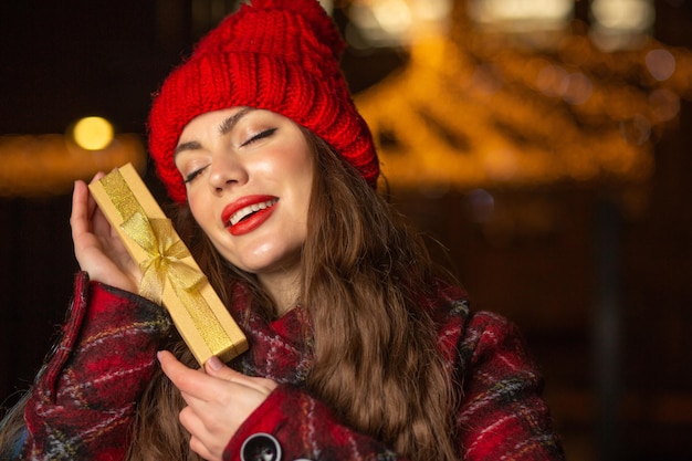 Delightful young woman in red hat holding gift box at the Christmas fair in the evening. Space for text