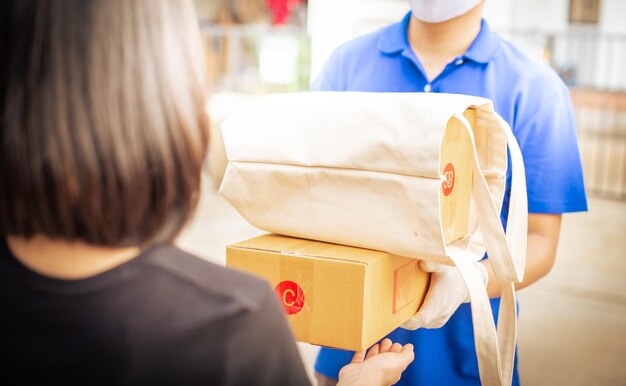 Deliver man wearing face mask in blue uniform handling bag and parcel box give to female costumer