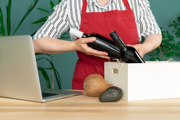 Deliver in a red apron Puts Wine Bottles in to white Box with coconuts and avocado, online order concept.