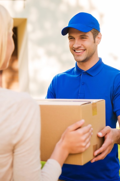 Delivering each package with smile. Happy young courier giving a cardboard box to young woman while standing outdoors