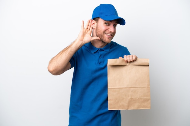 Delivery caucasian man isolated on white background listening to something by putting hand on the ear