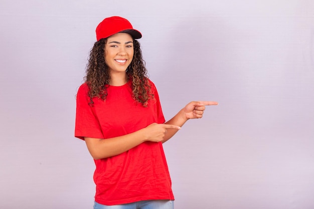 Delivery employee woman in red cap blank t-shirt uniform work courier in service during quarantine coronavirus covid-19 virus, pointing fingers aside on workspace isolated on white background studio