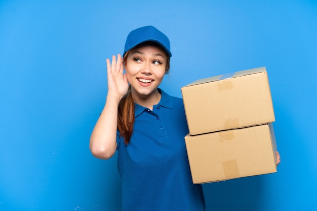 Delivery girl over isolated blue wall listening to something by putting hand on the ear