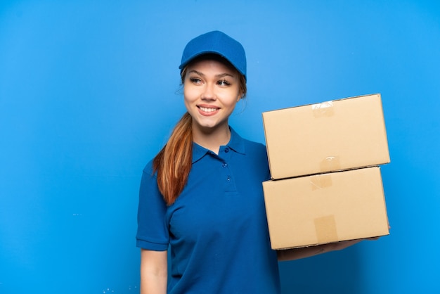 Delivery girl over isolated blue wall looking to the side and smiling