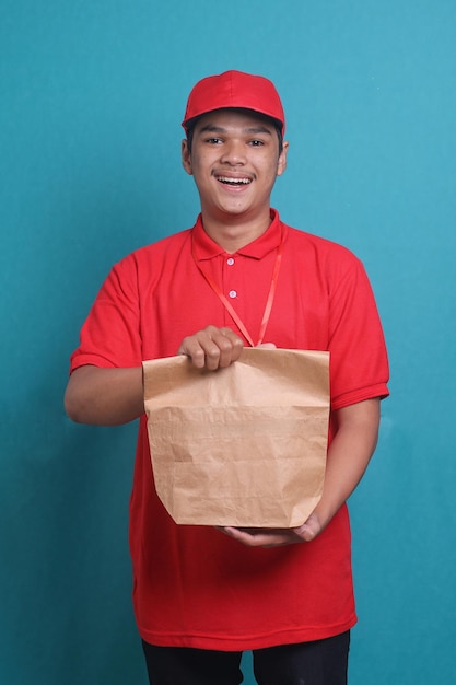 Delivery guy in red uniform holding craft paper bag from restaurant in blue studio background