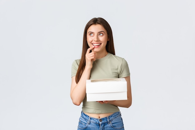 Delivery, lifestyle and food concept. Dreamy happy stylish woman looking up thoughtful, smiling as imaging something, holding box with desserts, delicious food order, stand white background.