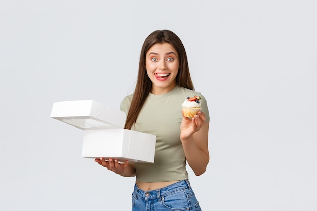 Consegna, stile di vita e concetto di cibo. una giovane donna sorridente eccitata ha ordinato una scatola con dolci dal caffè o dalla pasticceria preferiti, tenendo in mano un delizioso cupcake, leccandosi le labbra dal desiderio.