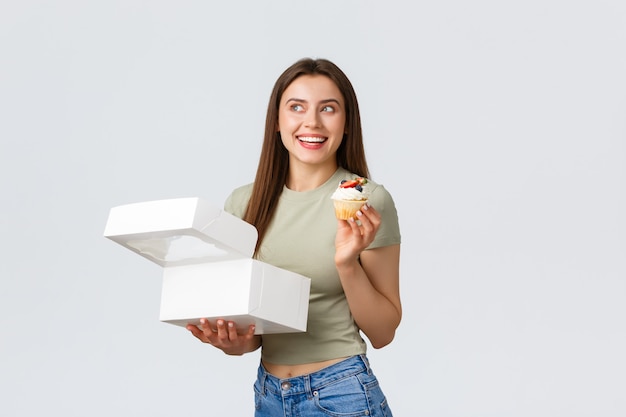 Concetto di consegna, stile di vita e cibo. bella donna adorabile che gode mangiando dessert dal caffè o dalla pasticceria. la ragazza ha ordinato sorridere dei bigné compiaciuto come come alimento dolce, fondo bianco