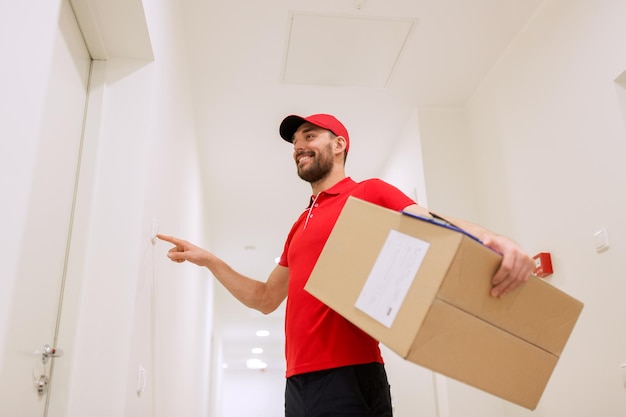配達、郵便、人、出荷のコンセプト – ドアベルを鳴らしながら廊下に宅配ボックスを持つ赤い制服を着た幸せな男