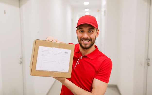 Foto concetto di consegna, posta, persone e spedizione - uomo felice in uniforme rossa con cassetta dei pacchi nel corridoio