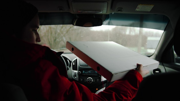 A delivery man drives a car and holding a pizza box