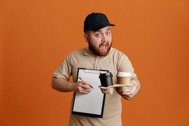 Dipendente dell'uomo delle consegne in berretto nero e uniforme della maglietta vuota che tiene tazze di caffè e appunti con la penna in attesa di una firma che sembra sorpresa e felice in piedi su sfondo arancione