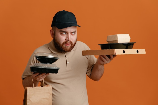 Delivery man employee in black cap and blank tshirt uniform holding food containers with paper bag looking aside with skeptic expression standing over orange background