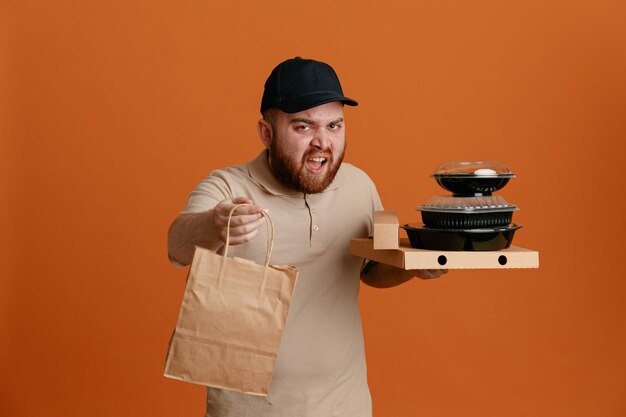 Delivery man employee in black cap and blank tshirt uniform holding food containers with paper bag looking at camera angry and frustrated shouting and yelling standing over orange background